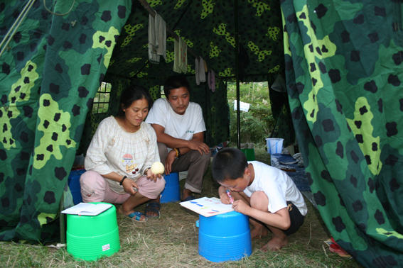 5月30日下午,九岁的成鸿钢(现就读九洲帐篷学校三一班)在防震棚门口作画。母亲罗财富和父亲成发志很喜欢看儿子作画,但他们在评价儿子的作品方面意见不一。母亲认为儿子用色不太合理,父亲则认为儿子色彩用得很好。中国文联网
